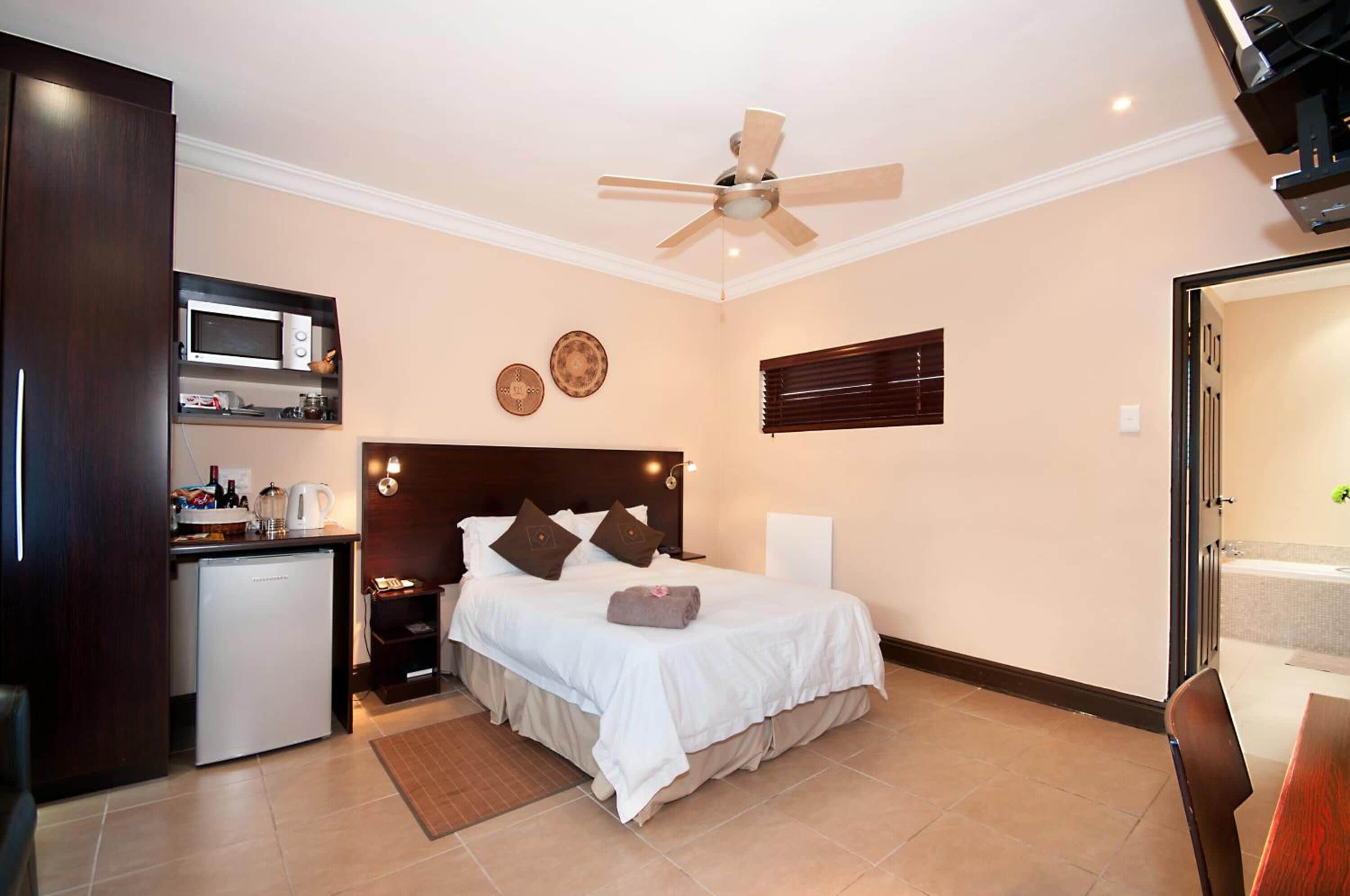 A compact hotel room with a bed, a small kitchenette, a ceiling fan, and a view of the outside.