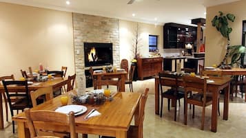 A spacious dining area with wooden tables and chairs, a stone fireplace, and a modern kitchen in the background.