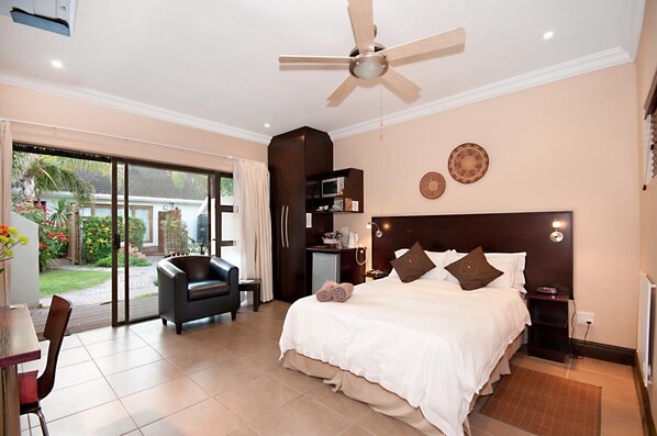 A hotel room with a bed, a desk, a chair, a ceiling fan, and a view of the outdoors.