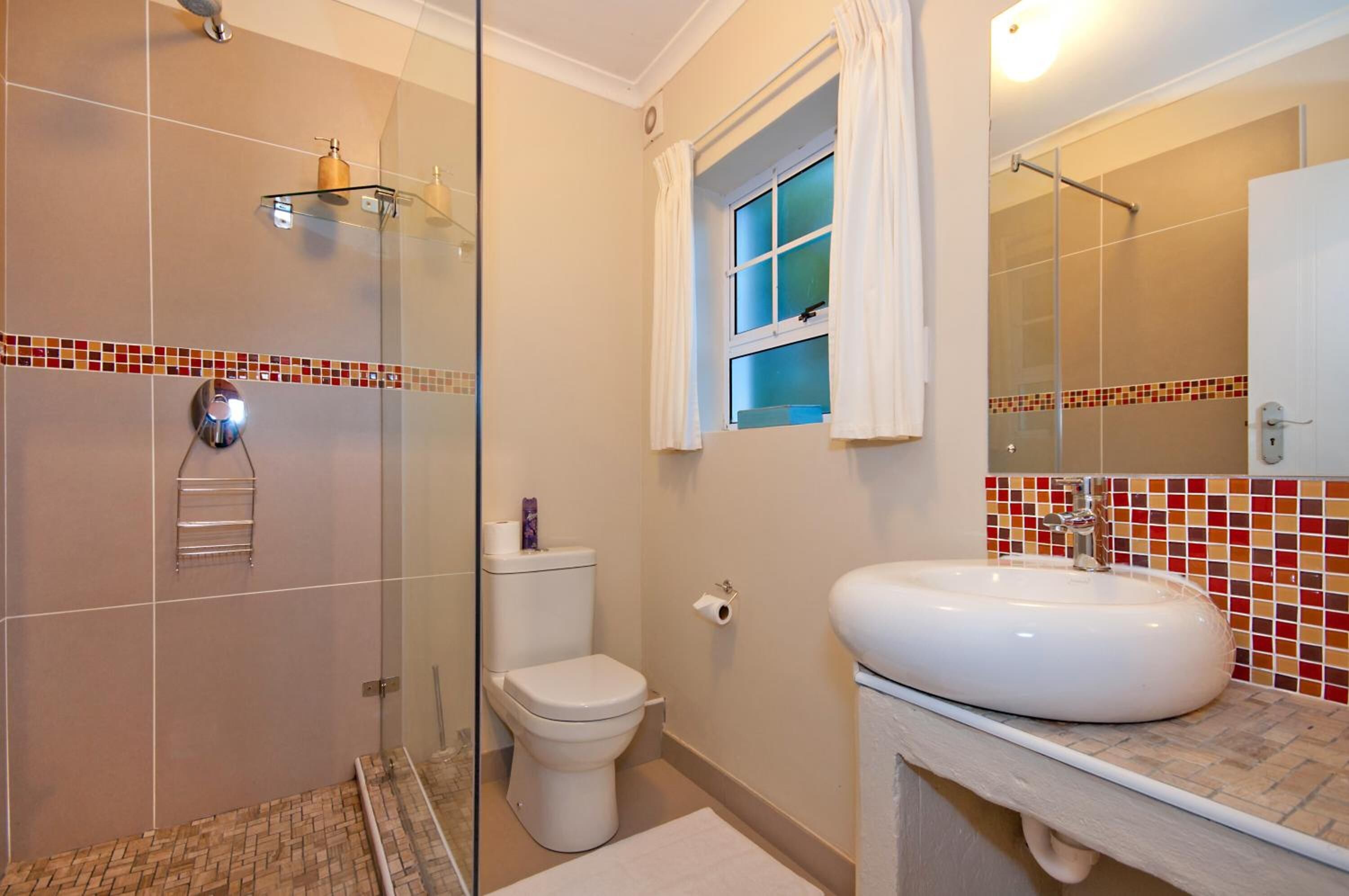 A bathroom with a glass shower enclosure, a toilet, a sink with a mirror, and a window with curtains.