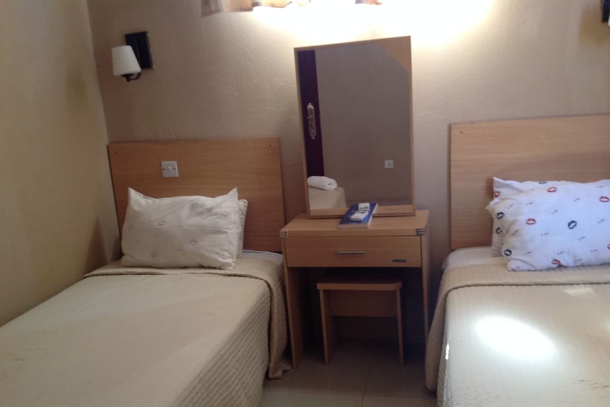 A small hotel room with two single beds, a wooden nightstand with a mirror, and a small desk with a chair.