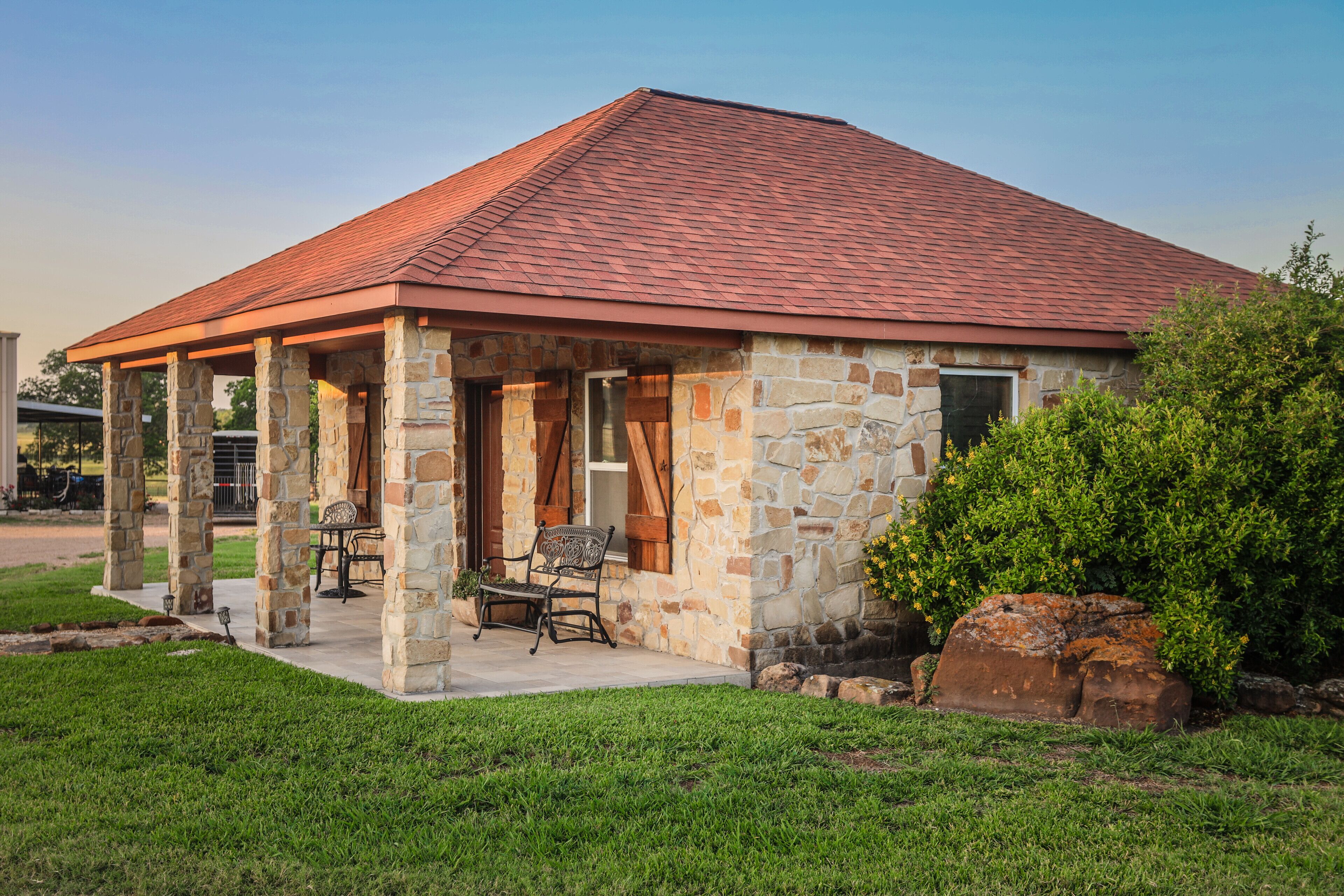 A stone building with a red roof, a covered patio with two chairs, and a large rock in the foreground.