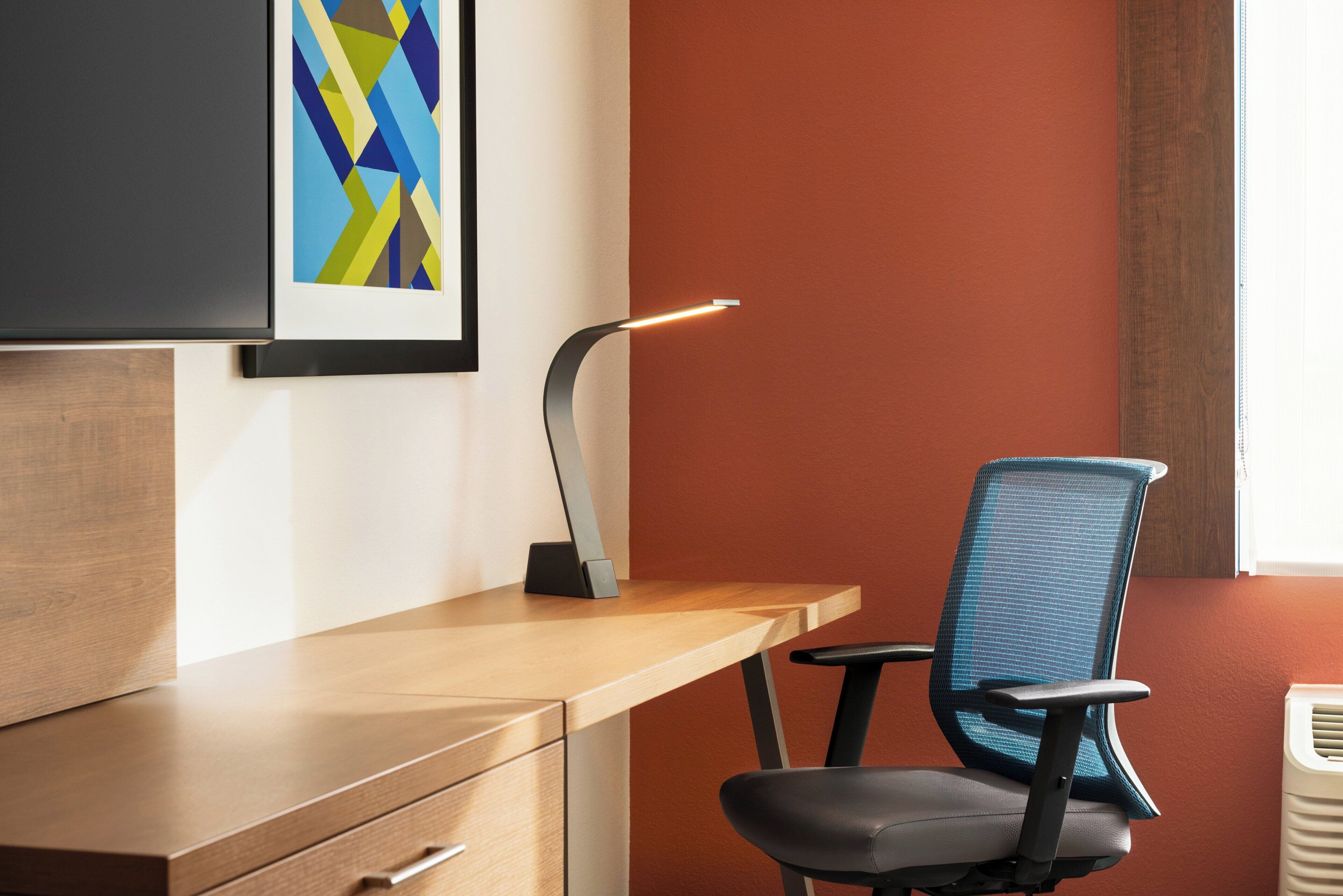 A modern office desk with a chair, a desk lamp, and a framed abstract artwork on the wall.