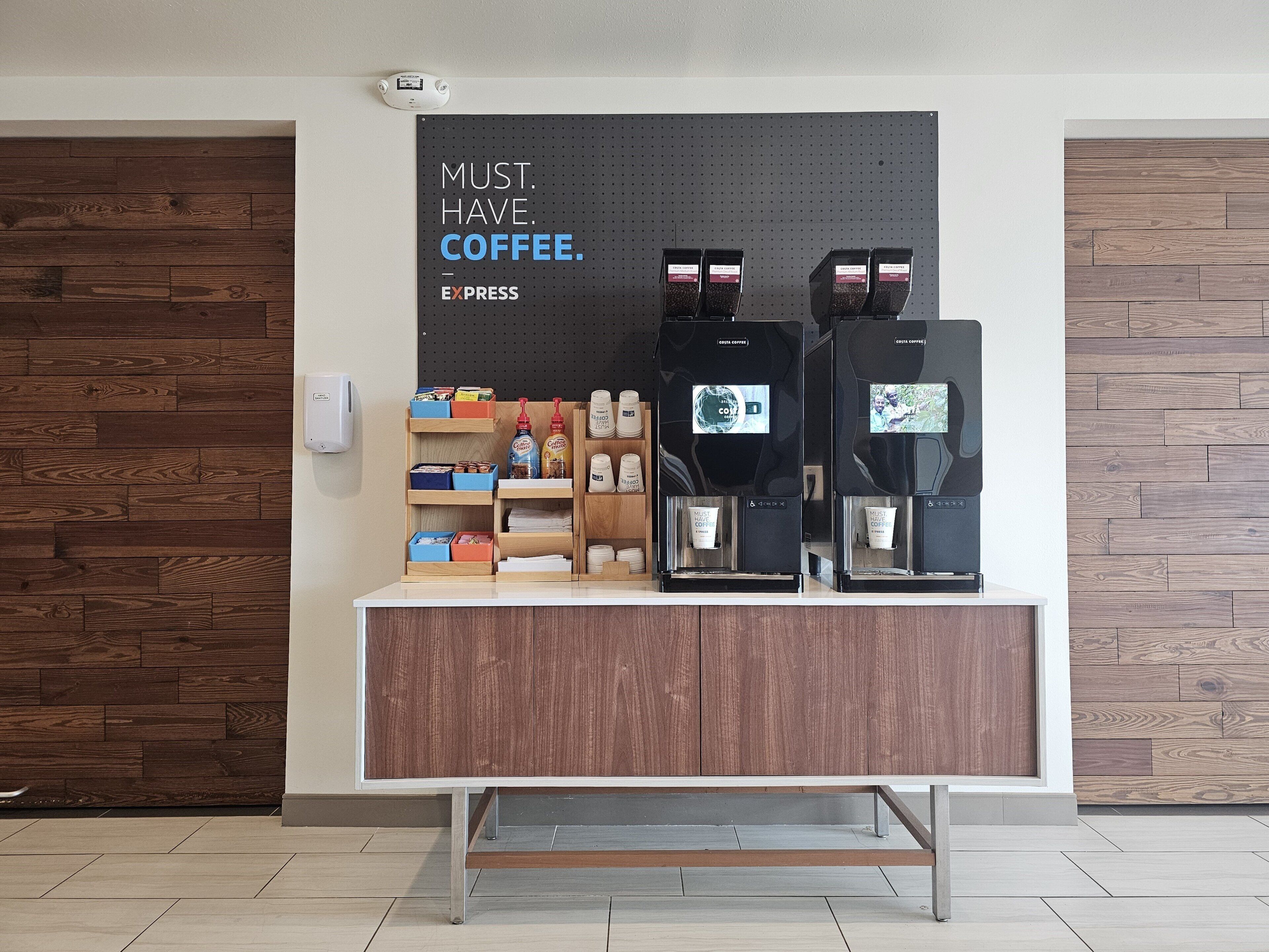 A coffee station with two espresso machines, a coffee dispenser, and a sign that reads 'MUST. HAVE. COFFEE.'