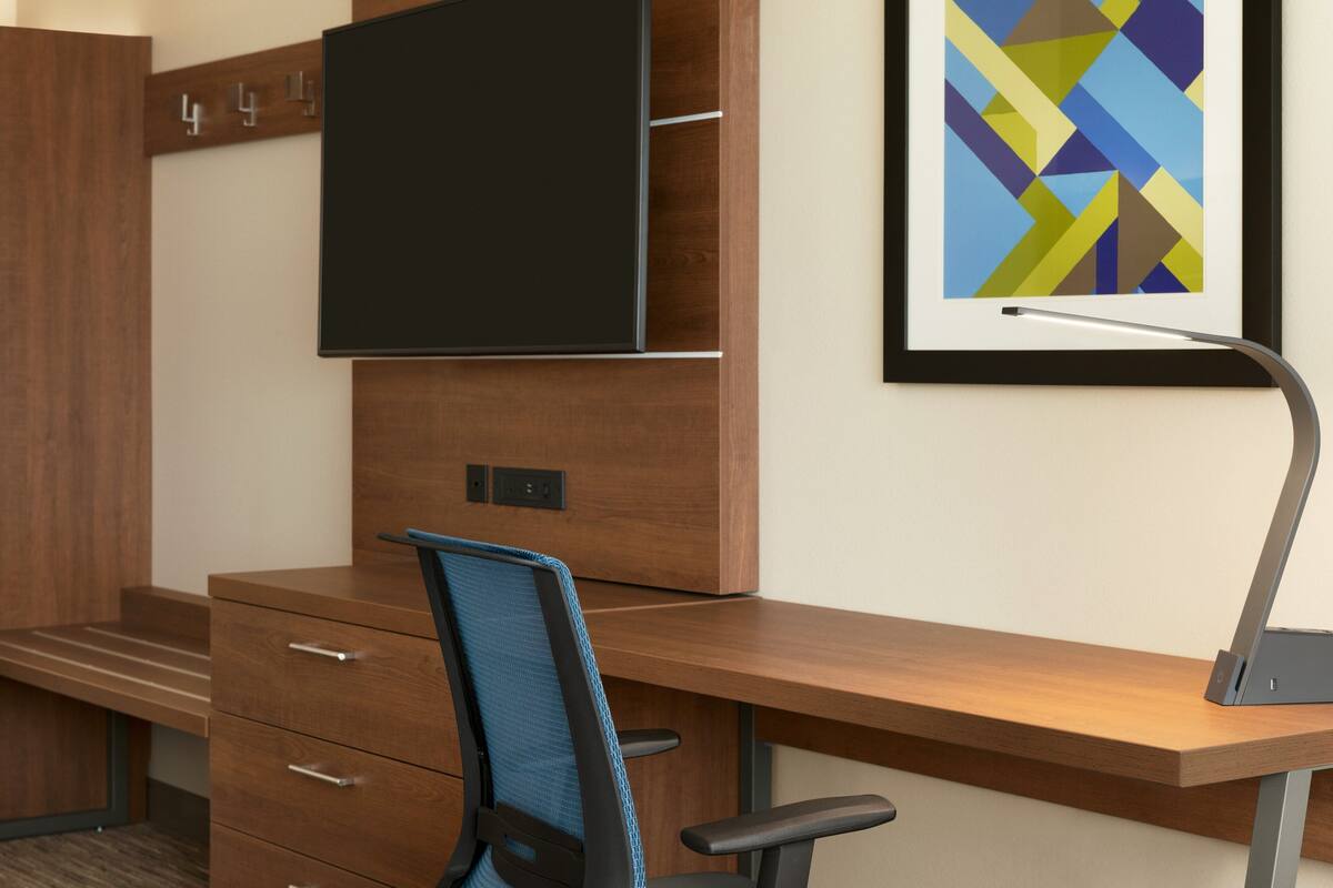 A modern office space with a wooden desk, a blue office chair, a mounted flat-screen TV, and a framed abstract art piece on the wall.
