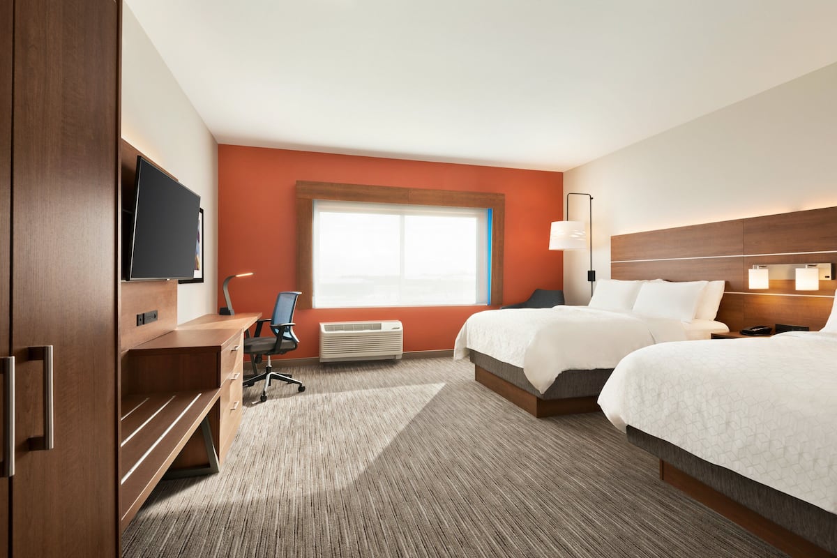 A hotel room with a wooden desk, a flat-screen TV, a large window, two beds, a nightstand, and a lamp.