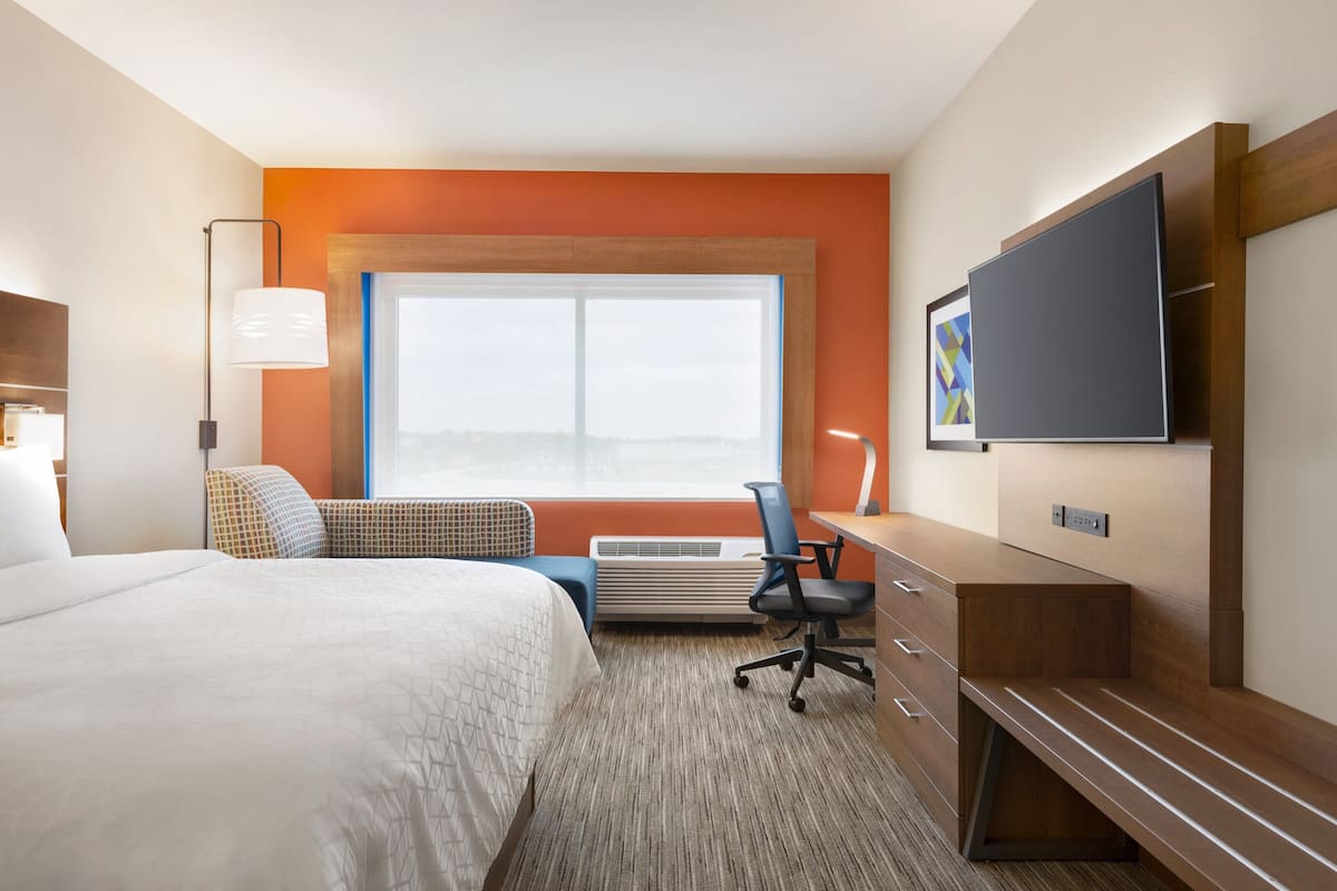 A hotel room with a large bed, a desk with a chair, a flat-screen TV, and a window with a city view.