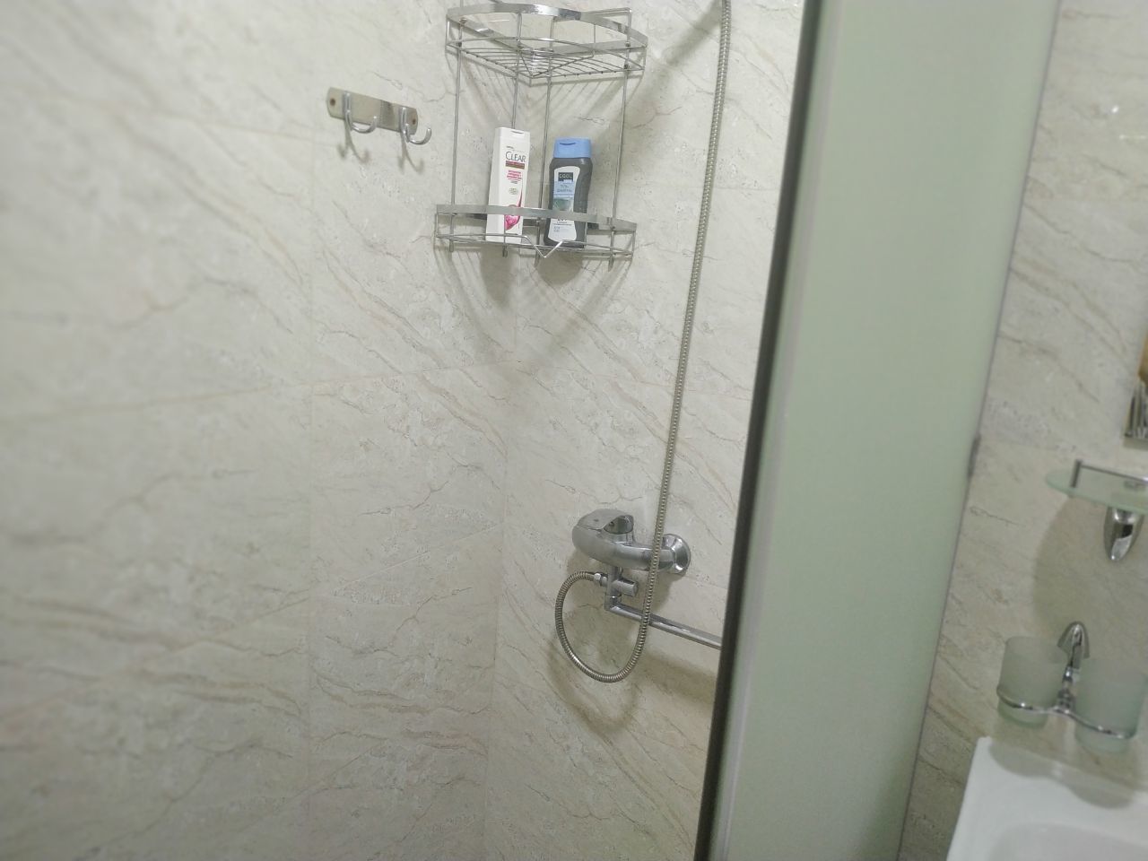Un espace douche doté d’une porte en verre, d’un pommeau de douche en métal et d’un distributeur de savon placé sur une étagère.