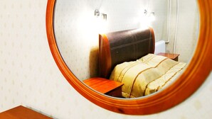 En rund spegel med en träram som reflekterar ett sovrum. Rummet har en mönstrad tapet, en brun läderklädd sänggavel och ett nattduksbord med en lampa.