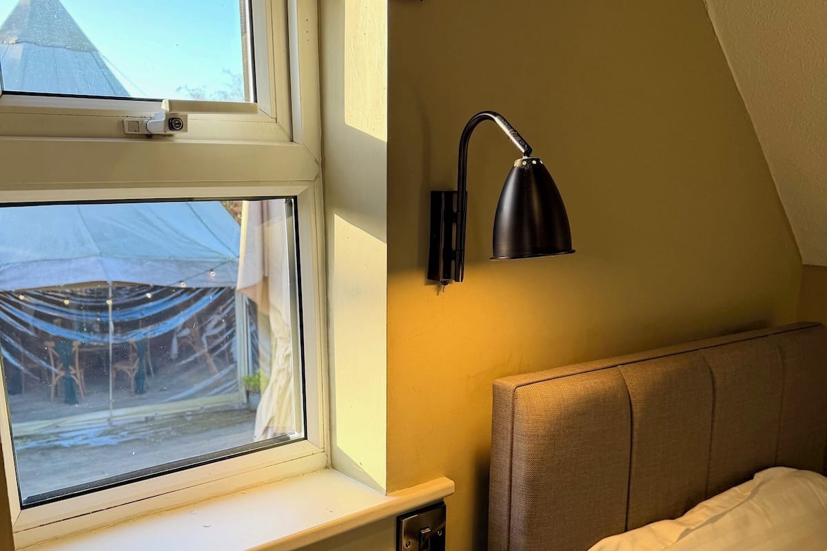 Une chambre avec une fenêtre, une lampe de chevet, un lit avec une literie blanche et une petite table avec un verre.