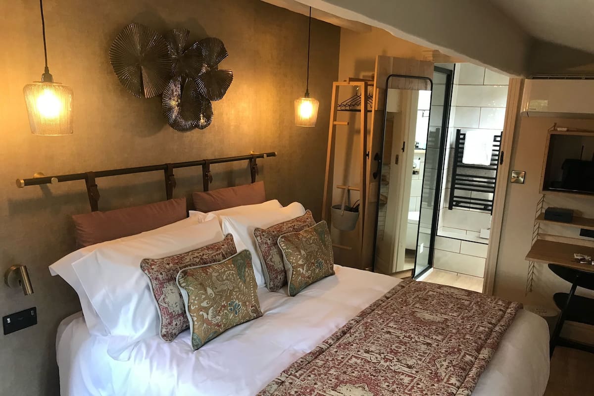 Un lit bien fait, agrémenté d’oreillers à motifs, un élément décoratif mural et un miroir qui reflète une salle de bain.