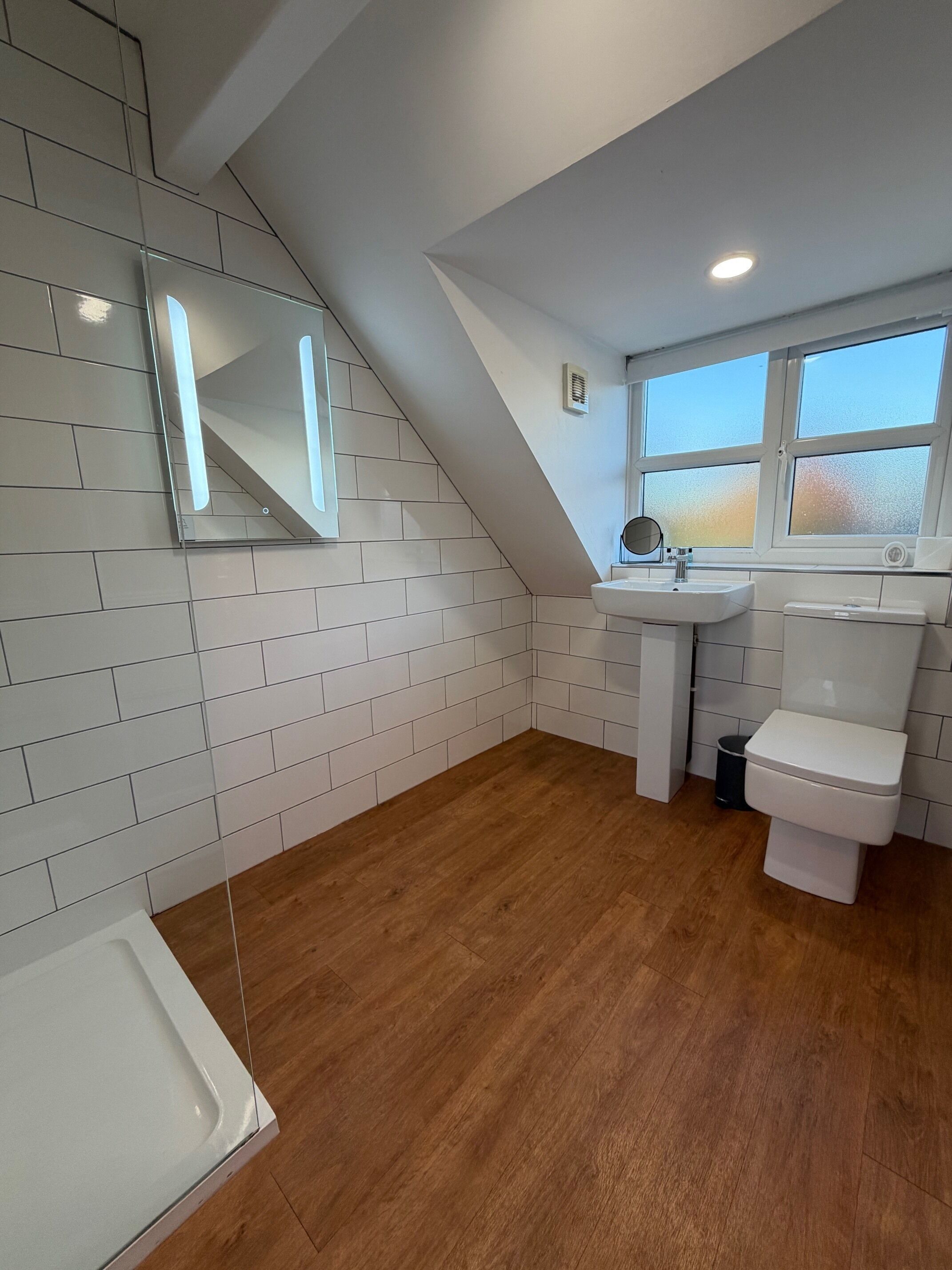 Une salle de bain moderne avec des murs recouverts de carreaux blancs, un plancher en bois, un lavabo blanc, des toilettes et un miroir au-dessus du lavabo.