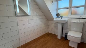 Une salle de bain moderne avec des murs recouverts de carreaux blancs, un plancher en bois, un lavabo blanc, des toilettes et un miroir au-dessus du lavabo.
