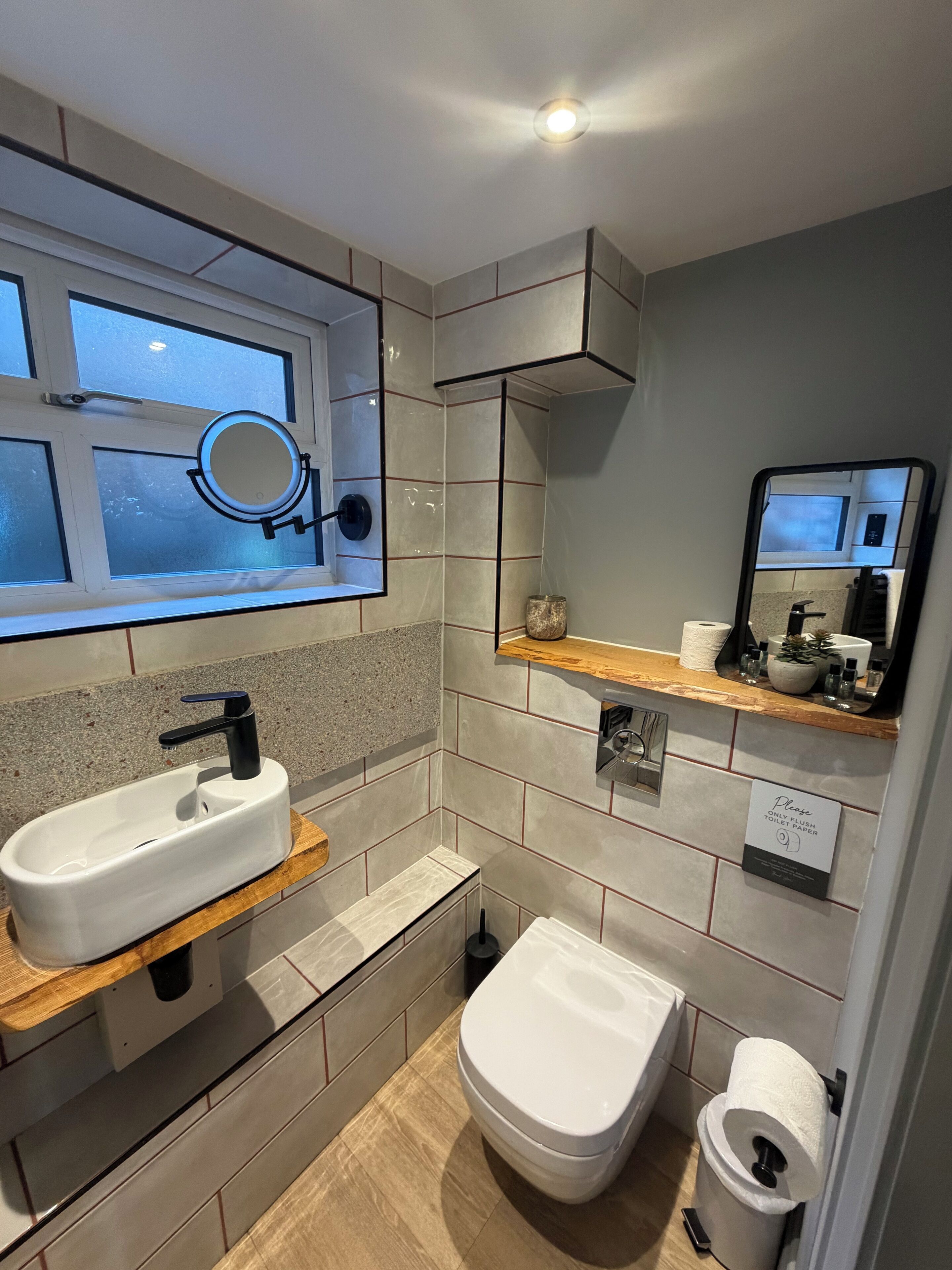 A modern bathroom with a white sink, black faucet, and a toilet. There is a mirror above the sink, a window with frosted glass, and a shelf with toiletries.
