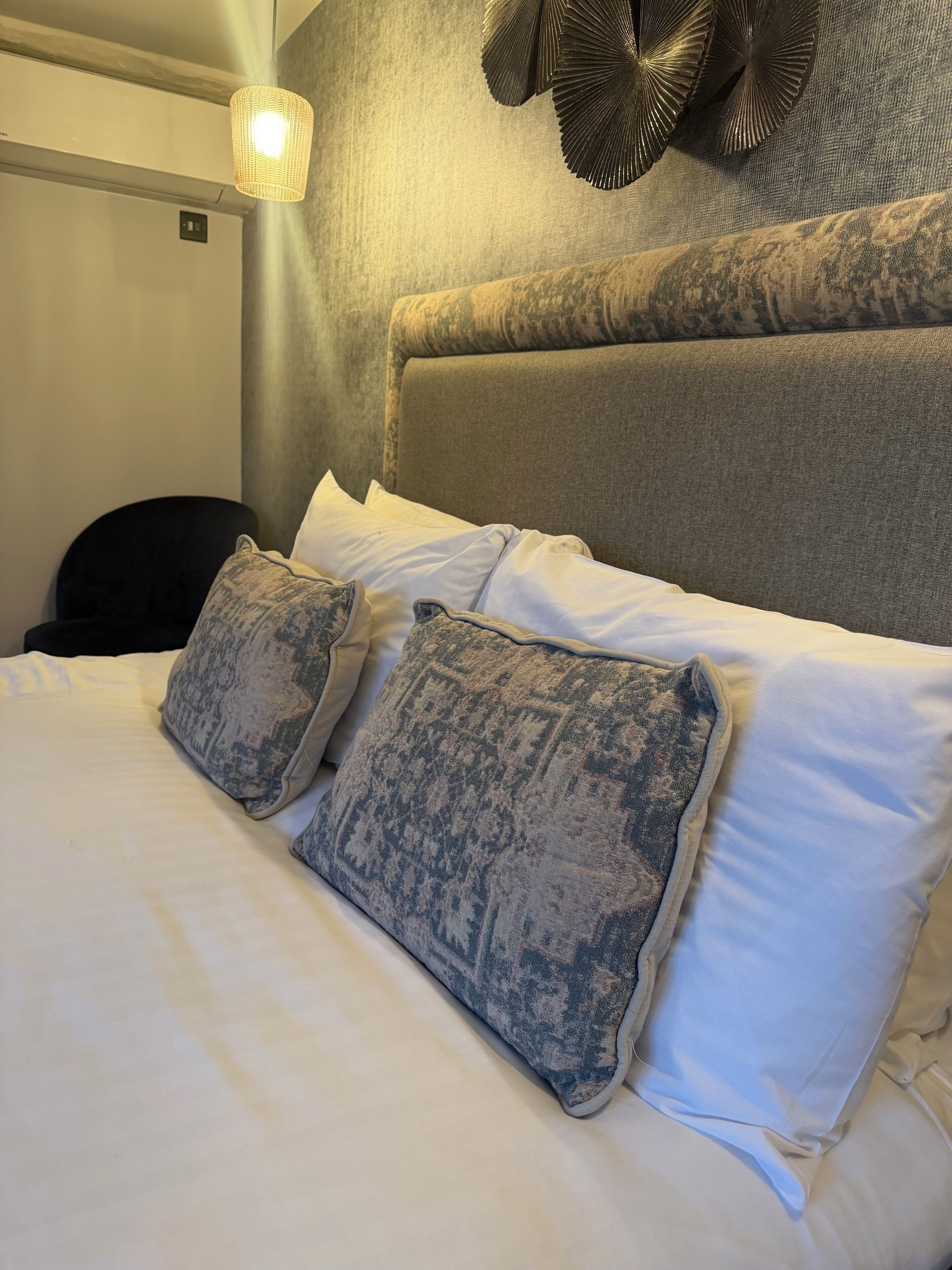 Una cama bien hecha con almohadas estampadas, un cabecero texturizado y una lámpara fijada a la pared.