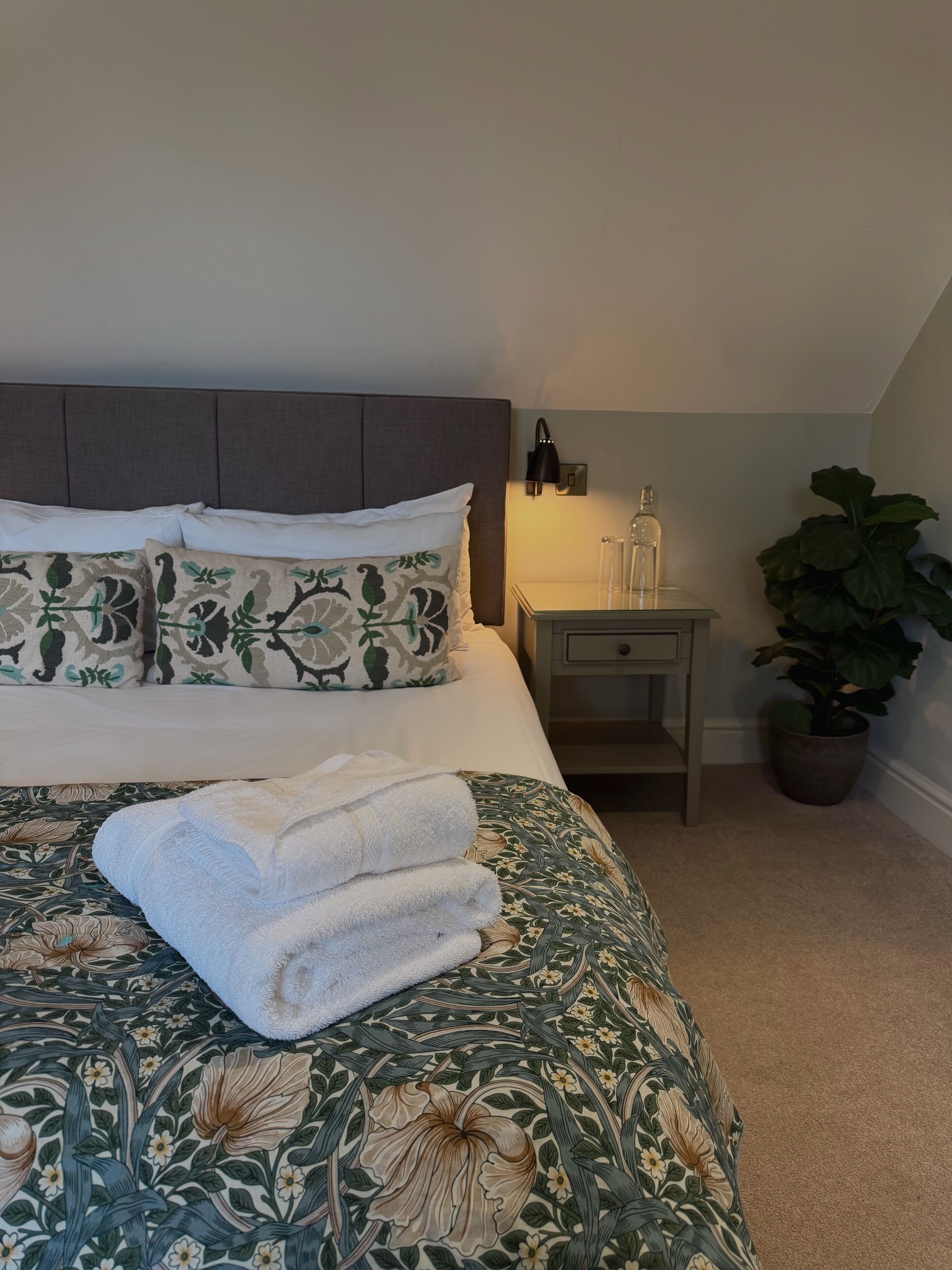 A neatly made bed with floral bedding, a bedside table with a lamp, and a potted plant.
