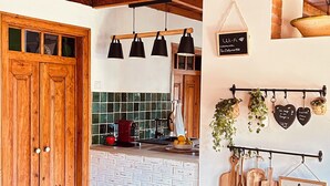 Una mesa de comedor de madera con sillas, una zona de cocina con salpicadero de azulejos, lámparas colgantes y un mueble de pared con utensilios de cocina.