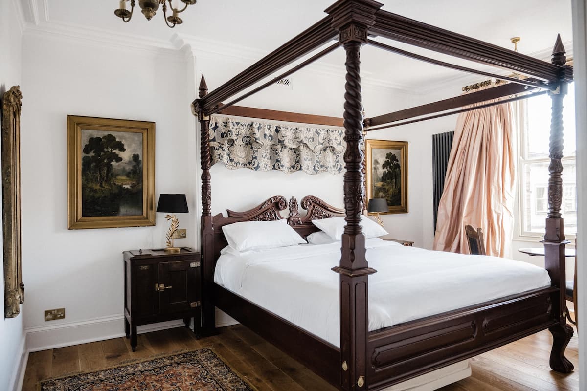 A four-poster bed with a dark wooden frame, a white bedspread, and matching pillows. There is a nightstand with a lamp and a framed picture on the wall to the left.