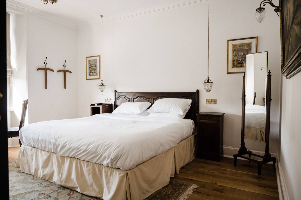 A neatly made bed in a room with a wooden floor, a bedside table, a nightstand, a mirror, and wall-mounted lights.