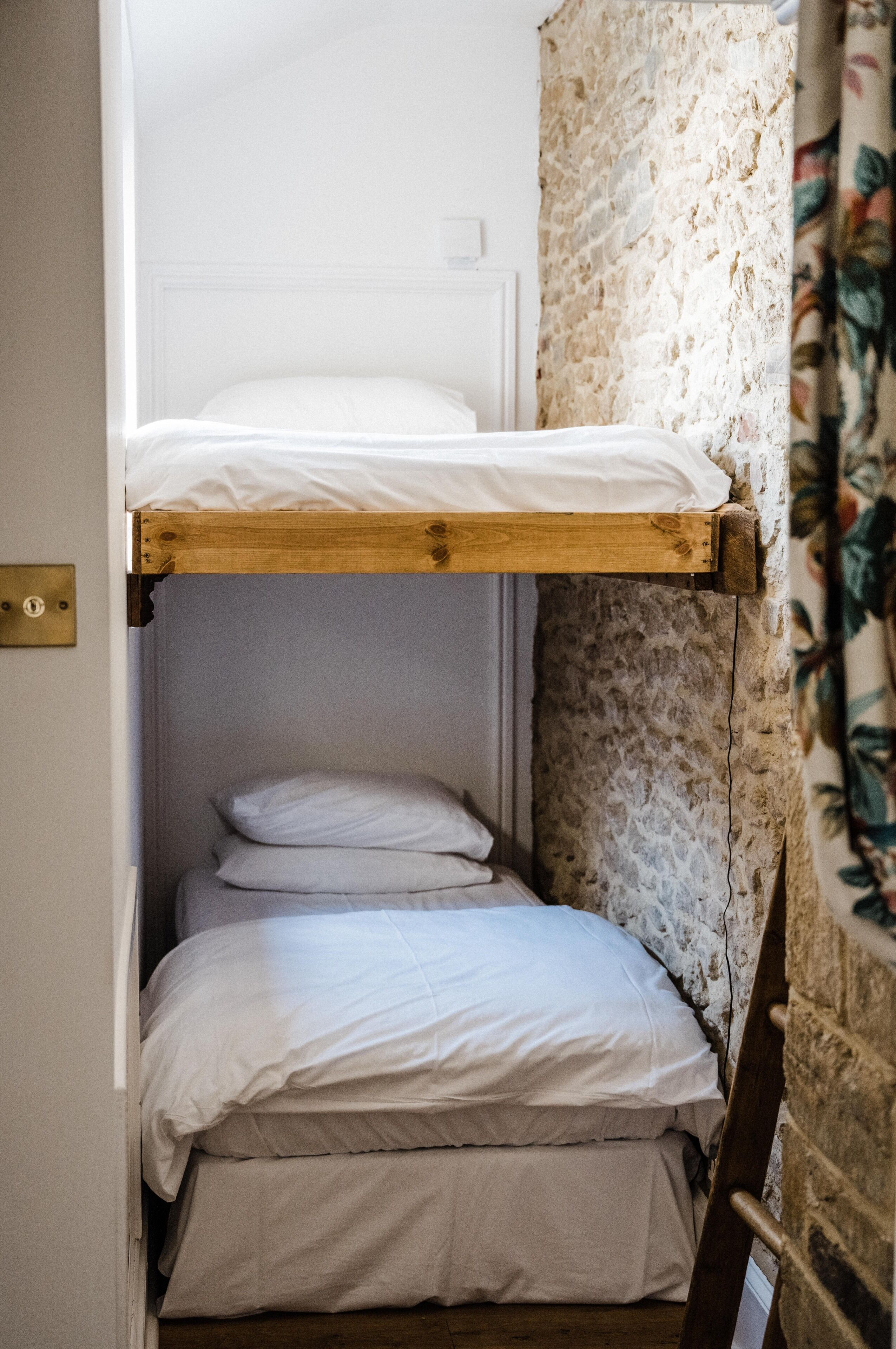 Een stapelbed met witte beddengoed in een kamer met stenen muren en een bloemen gordijn.