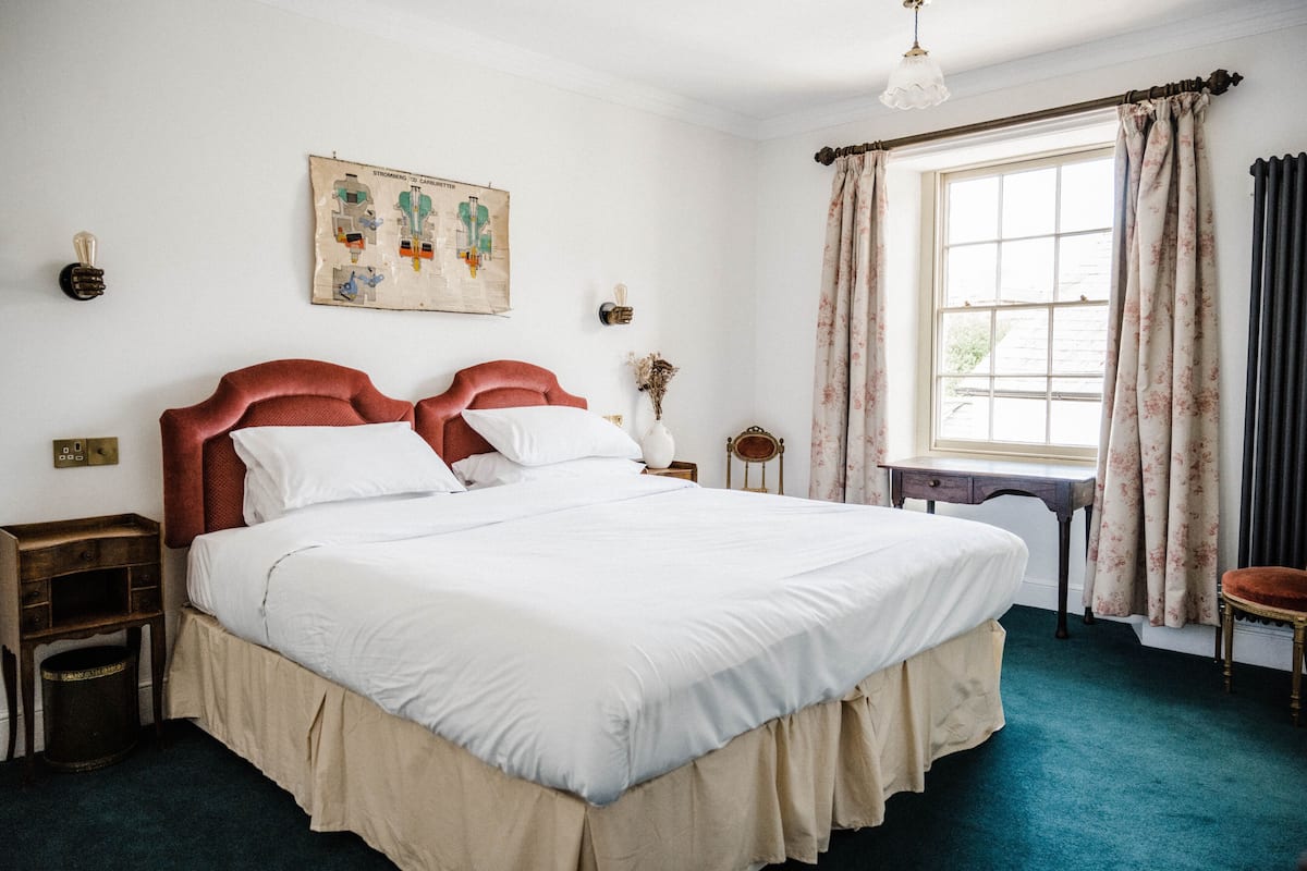 A large bed with a headboard, a nightstand, a small table, a chair, a window with curtains, and a wall-mounted light fixture.