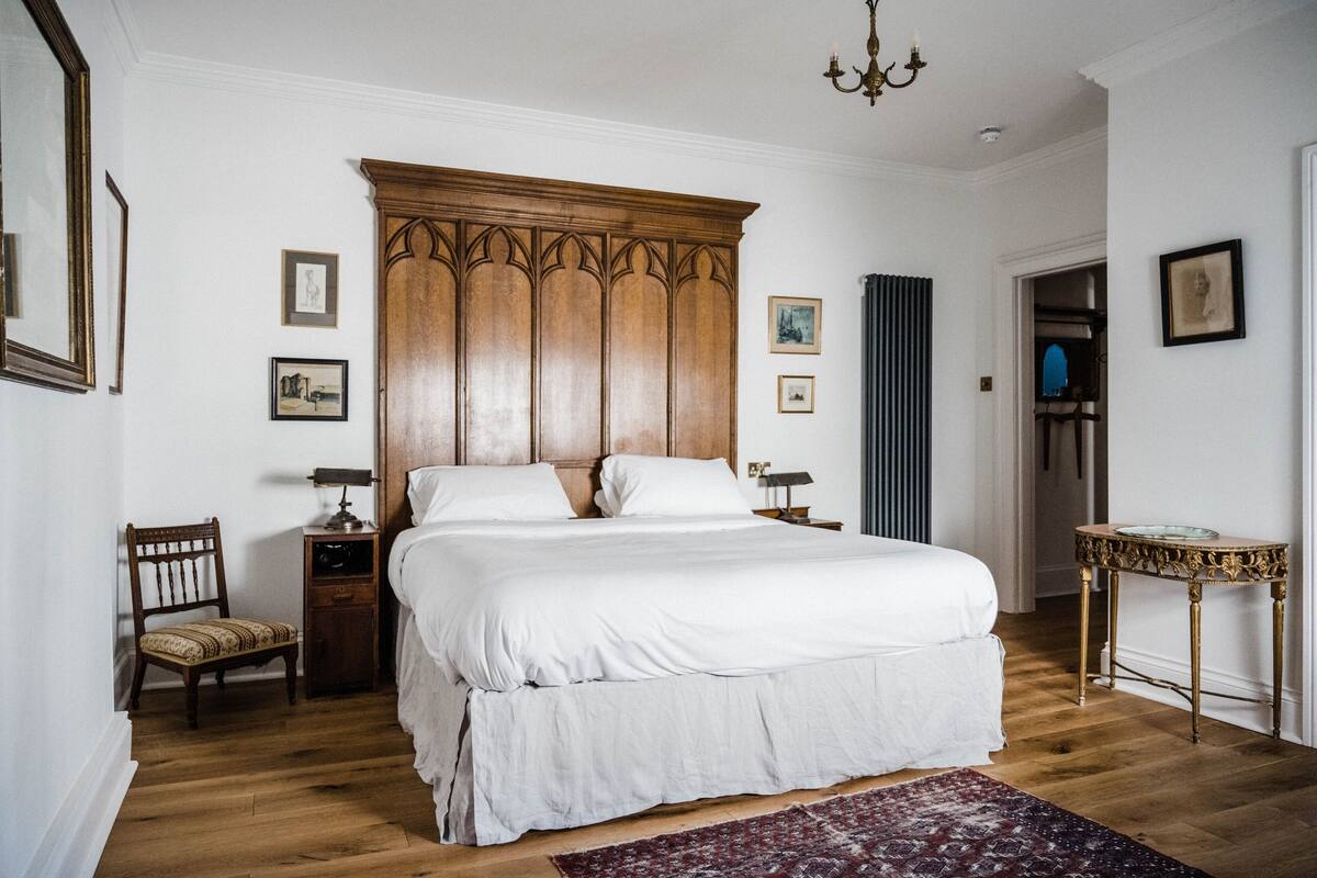 A bedroom with a large wooden bed, a chair, a small table, and framed pictures on the walls.