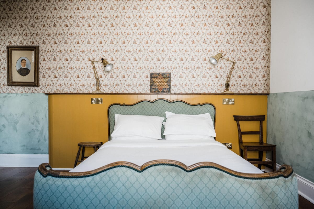 A bed with a patterned headboard, a framed portrait, and two wall-mounted lamps.
