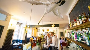 A bar area with a large bird sculpture, a variety of liquor bottles, and patrons. - The Crooked Swan (Crewkerne)