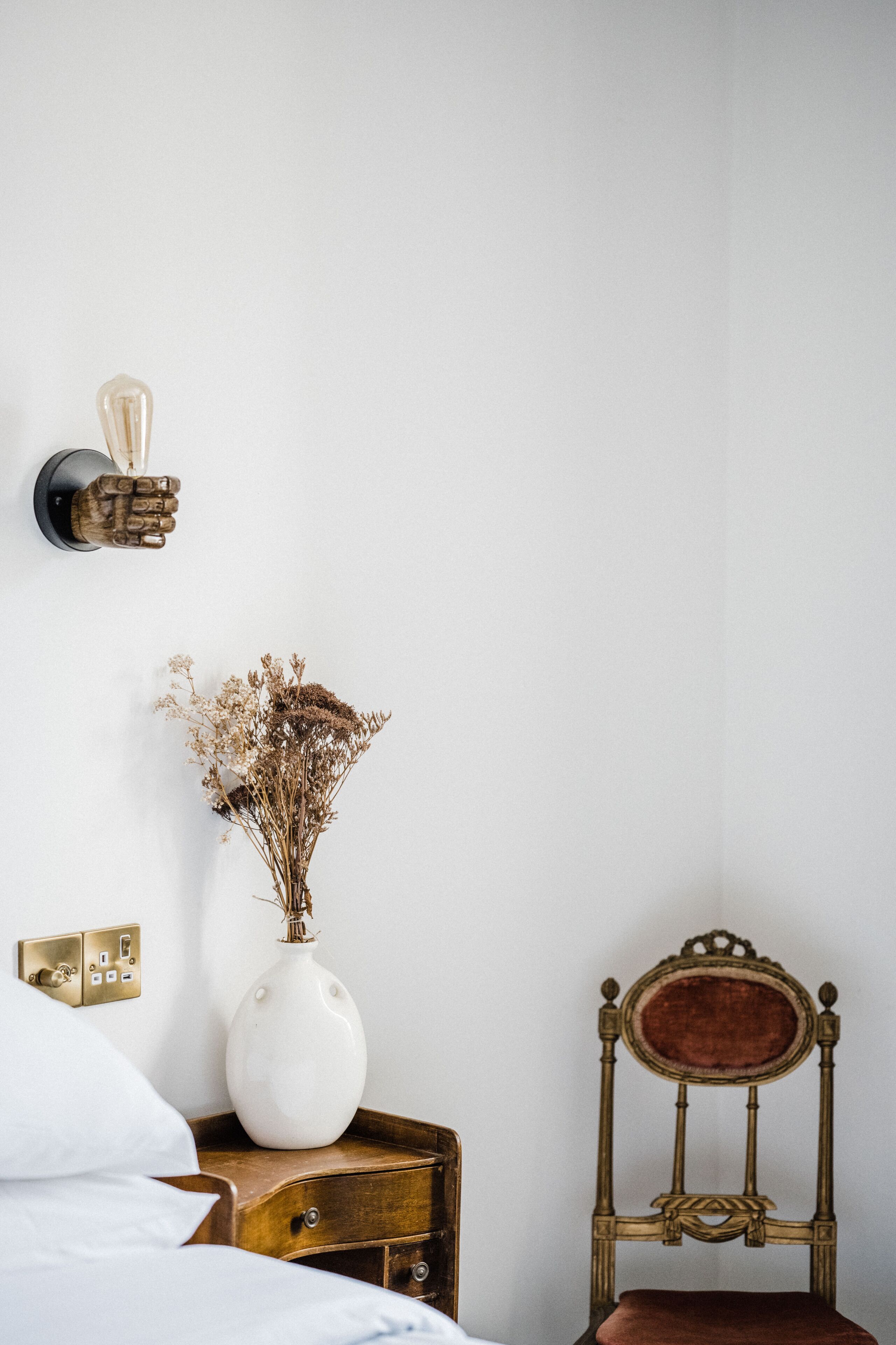 Un dormitorio con pared blanca, mesita de noche de madera, florero blanco con flores secas, una silla antigua y una lámpara de pared.