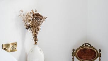 Un dormitorio con pared blanca, mesita de noche de madera, florero blanco con flores secas, una silla antigua y una lámpara de pared.