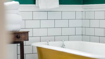 A bathroom with a white bathtub, a wooden side table with towels, and a green wall with a framed picture.