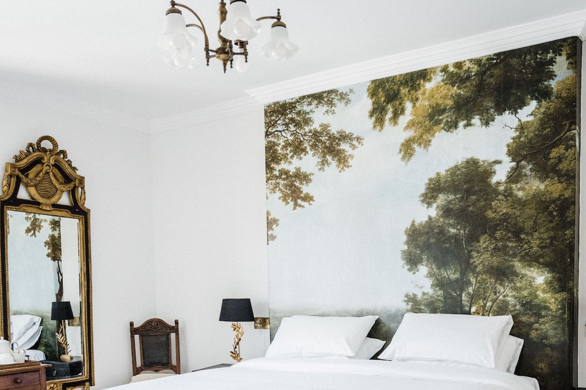 A large bed with white bedding, a chandelier, a large mirror, and a wall mural of trees.