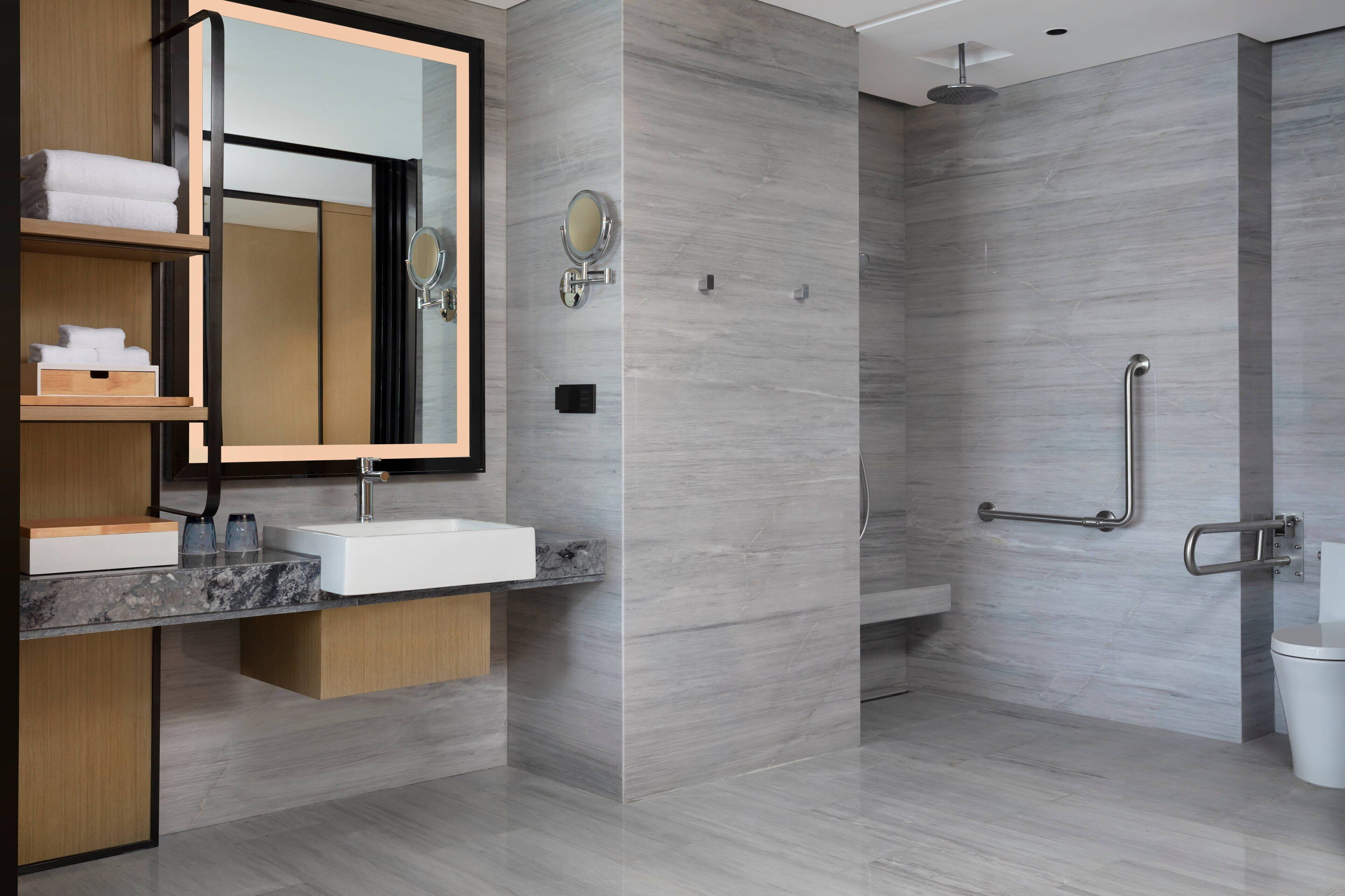 A modern bathroom with a marble sink, wooden shelves, a large mirror, and a walk-in shower with grab bars.