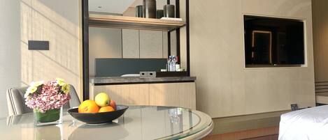 Una habitación de hotel moderna con una mesa de comedor de vidrio, un cuenco de frutas, un jarrón de flores, un espejo grande y un televisor.