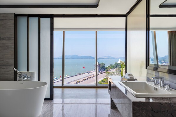 A modern bathroom with a large bathtub, a double sink, and a large window offering a view of a coastal area.