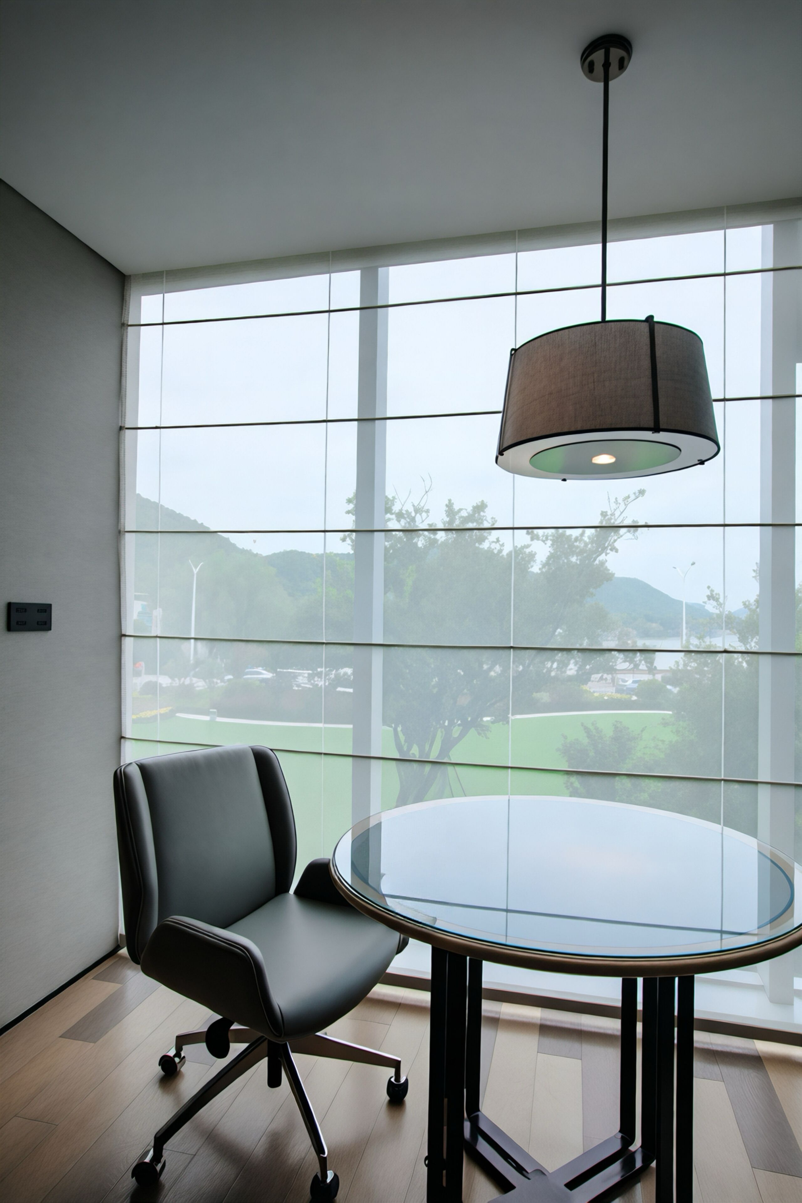 A modern office space with a round glass table, a black office chair, and a hanging lamp.