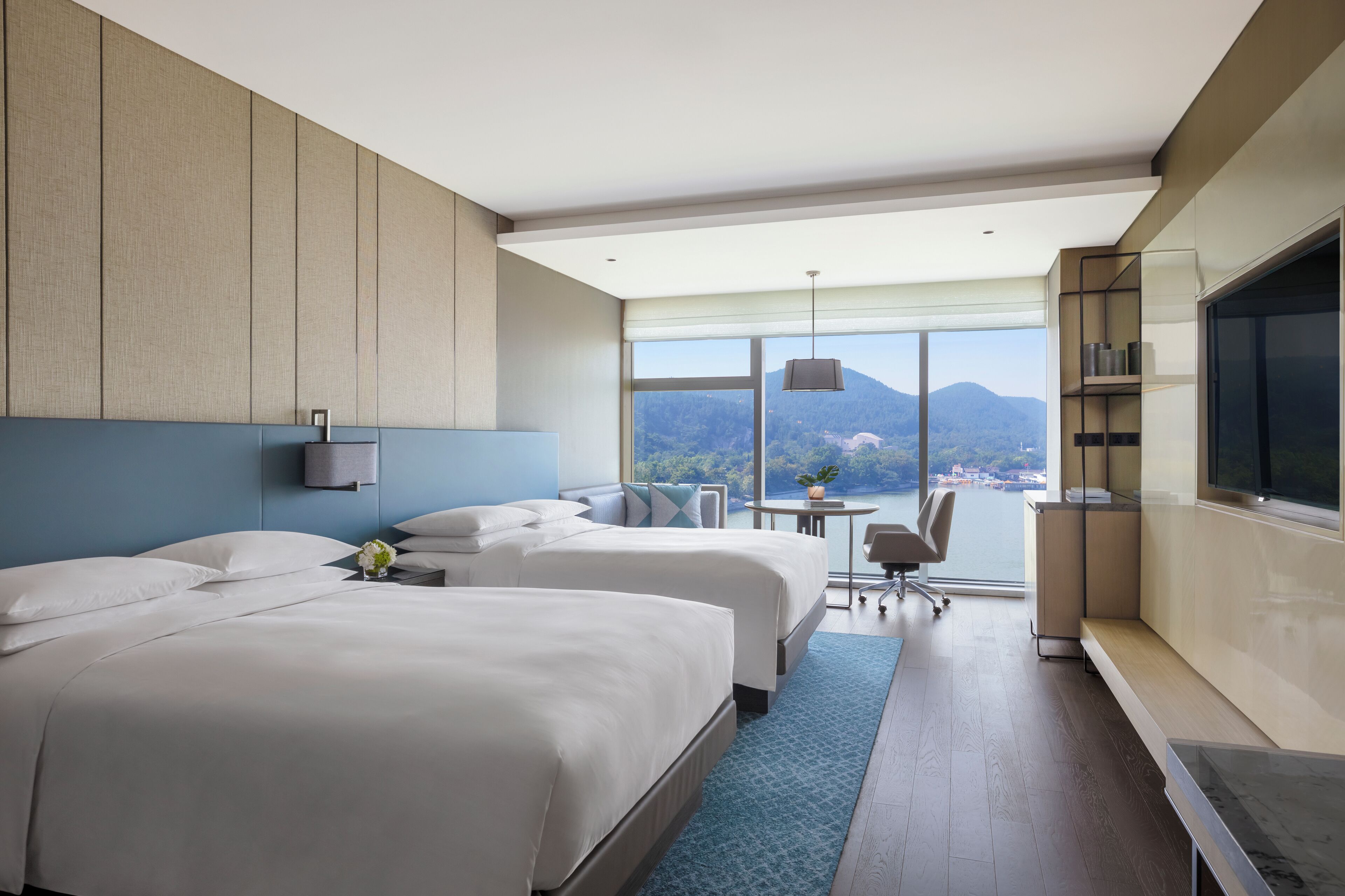 A hotel room with two beds, a desk, a chair, a TV, and a view of mountains.