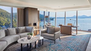 Una habitación de hotel moderna con un ventanal grande, un sofá, una silla, un escritorio y vistas a un lago y montañas.