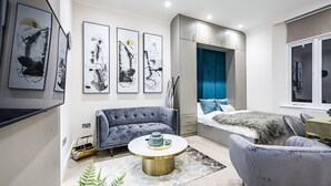 A modern living room with a grey sofa, a round coffee table, and a bed with blue bedding. There are abstract paintings on the wall and a window with blue curtains.