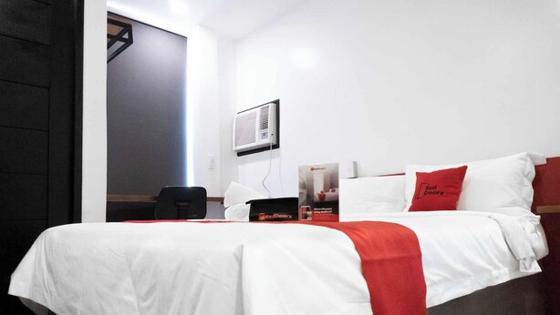 Een hotelkamer met een bed, een bureau, een stoel, een rood-wit kussen, een wandairconditioner en een deur.
