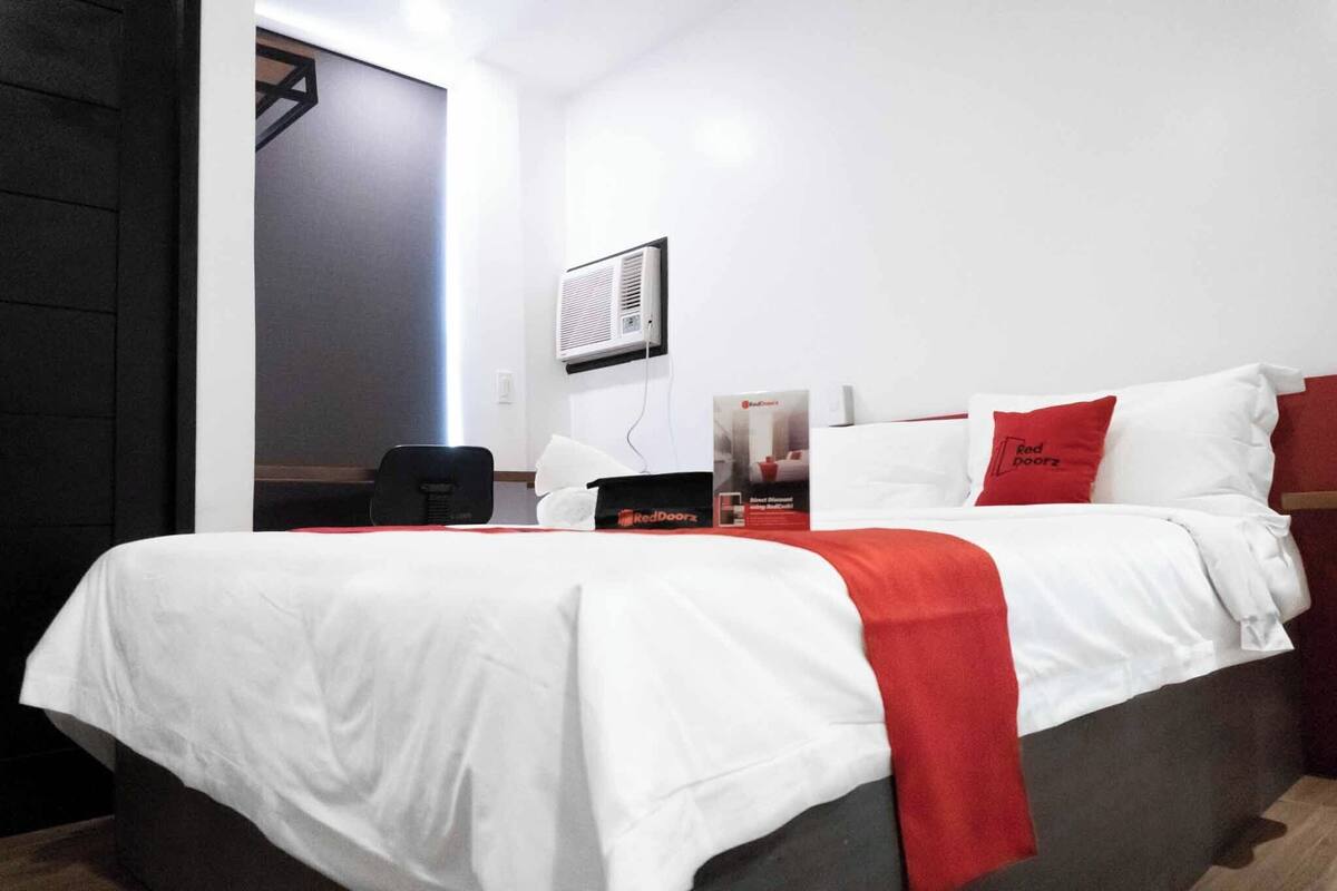 Een hotelkamer met een bed, een bureau, een stoel, een rood-wit kussen, een wandairconditioner en een deur.