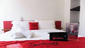 Een hotelkamer met een net opgemaakt bed, een kussenstel in rood en wit, een tas met het RedDoorz logo en een reclamezuil van RedDoorz.