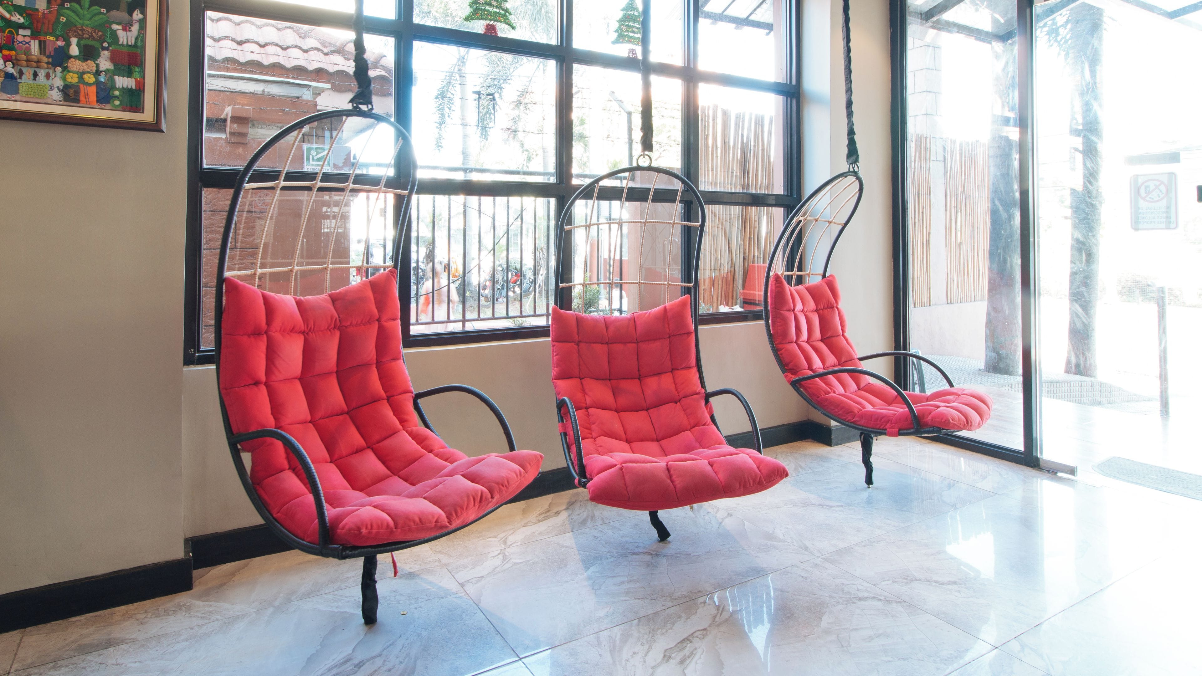 Three red cushioned chairs suspended from the ceiling in a room with large windows.