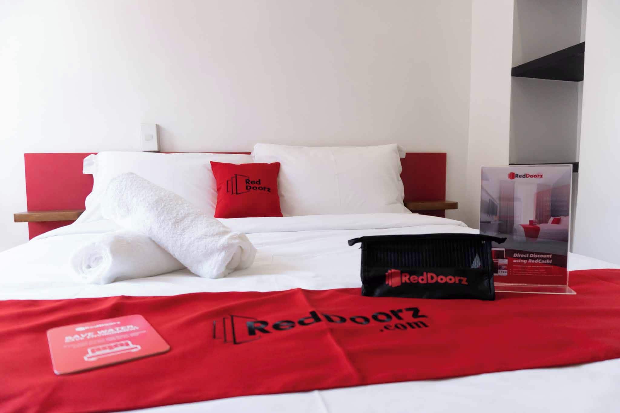 Ein Hotelzimmer mit einem ordentlich bezogenen Bett, einem roten und weißen Kissenpaar, einer Tasche von RedDoorz und einem Werbeständer von RedDoorz.