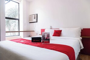 Ein Hotelzimmer mit Bett, einem Fenster mit Blick auf Palmen und einer Wand-Klimaanlage. Auf dem Bett ist ein RedDoorz-Schild zu sehen.