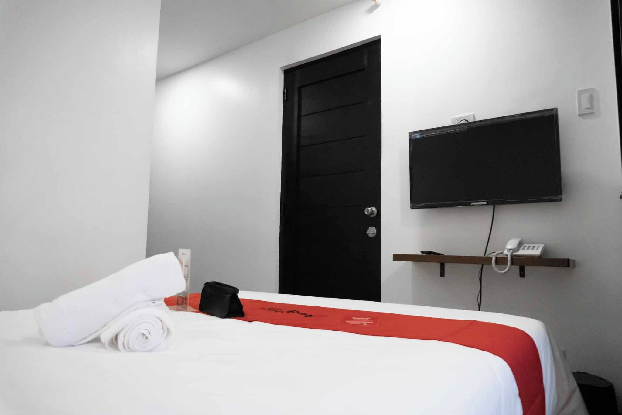 Una camera d'albergo con un letto, una porta nera, una TV a parete, un telefono su uno scaffale e un asciugamano bianco arrotolato sul letto.