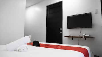 Una camera d'albergo con un letto, una porta nera, una TV a parete, un telefono su uno scaffale e un asciugamano bianco arrotolato sul letto.