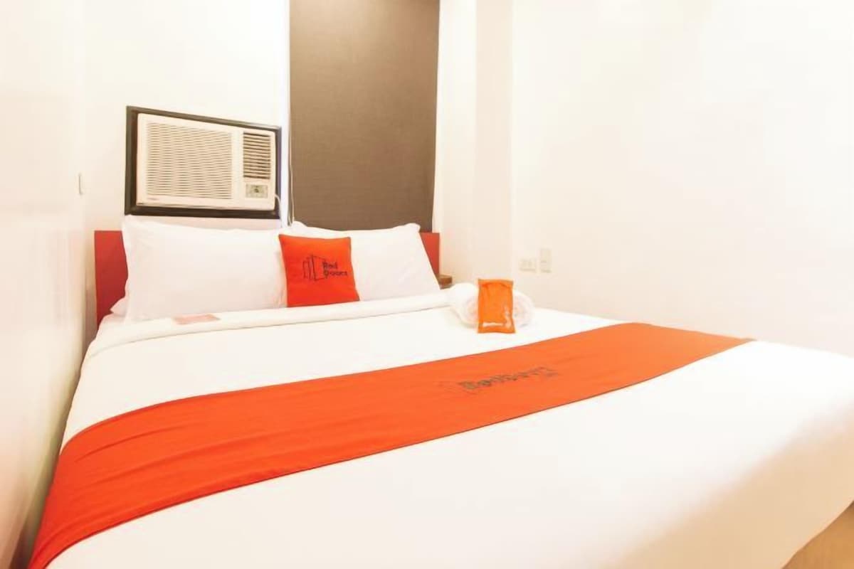 Ein Hotelzimmer mit einem Bett, einer roten und weißen Bettdecke, einem Kissen mit einem Logo und einer Wand-Klimaanlage.