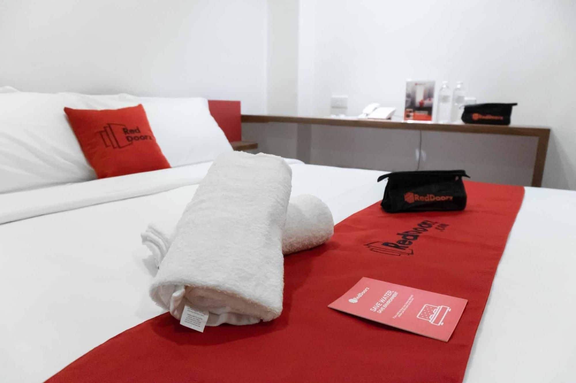 A neatly made bed with a white towel, a red pillow, and a red blanket with a RedDoor logo. There is a small black bag and a red card on the bed.
