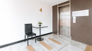 Een moderne lobby met een zwarte lederen fauteuil, een klein tafeltje met een plant, een marmeren vloer en een lift met een bordje erboven.